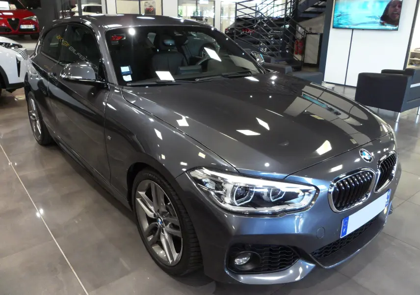 BMW Série 1 F21 LCI gris vue 3/4 avant droit, avec jantes alliage et phares LED allumés en showroom.
