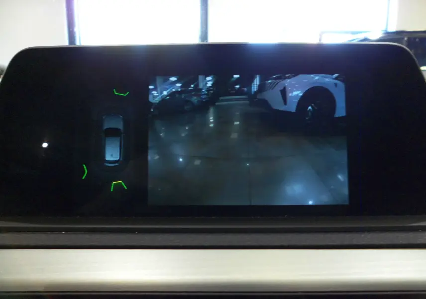 Écran de caméra de recul montrant un véhicule blanc en 3/4 arrière droit dans un parking, sur BMW Série 1 F21 LCI gris.