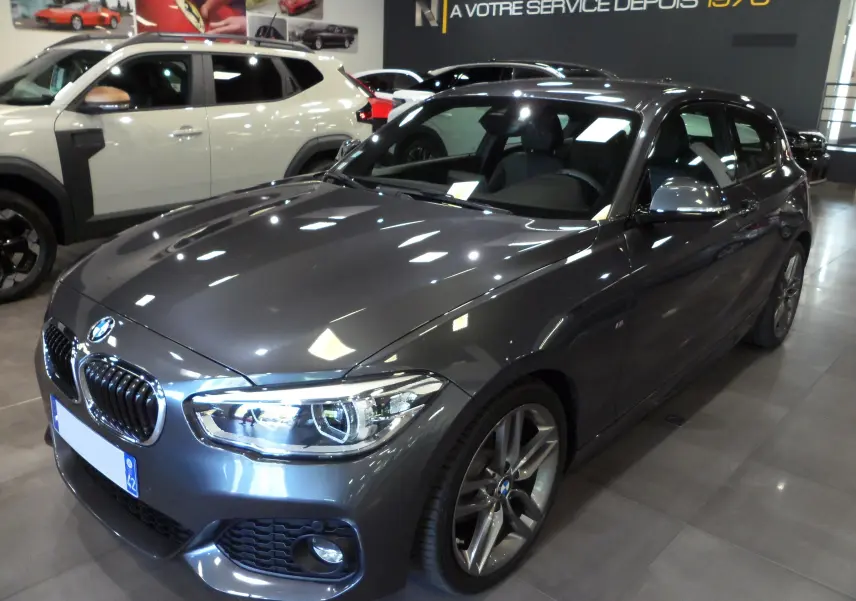 BMW Série 1 F21 LCI 118i gris vue 3/4 avant droit en intérieur, avec jantes alliage et phares LED allumés.