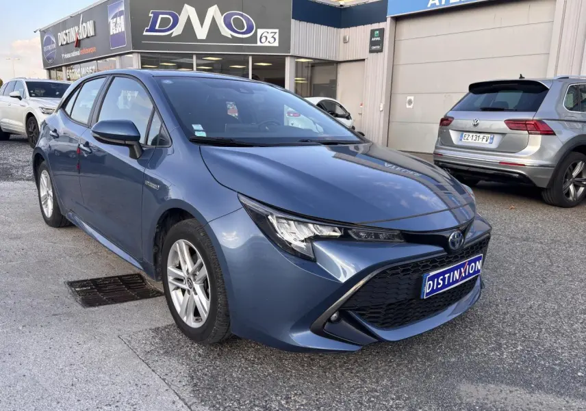 Toyota Corolla hybride bleu clair vue 3/4 avant droit, calandre noire et phares effilés, stationnée devant un atelier.
