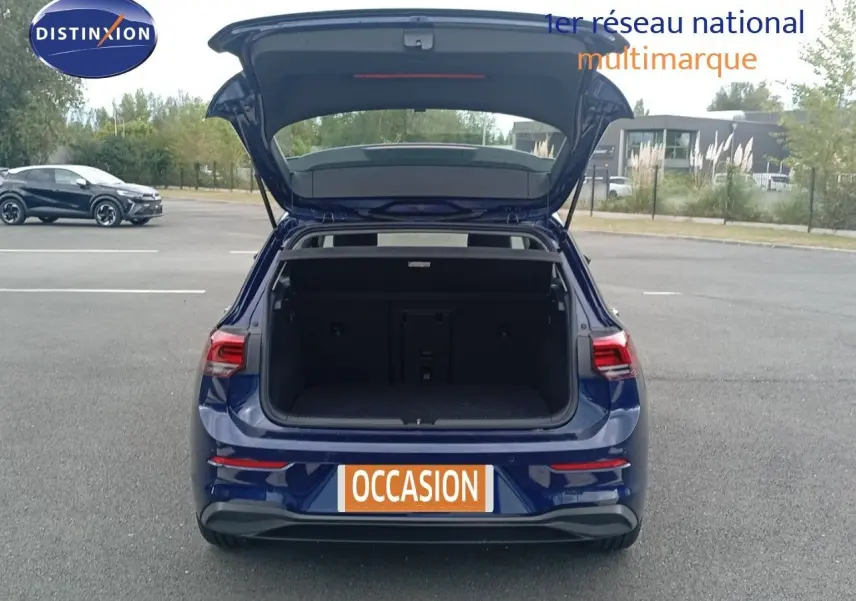 Vue arrière d'une Volkswagen Golf VIII bleu Atlantique métal avec coffre ouvert sur un parking extérieur.