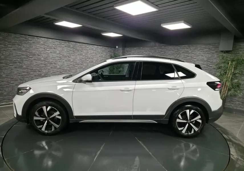 Profil droit du Volkswagen Taigo blanc 2024 avec jantes alliage 17 pouces et barres de toit noires dans un showroom.