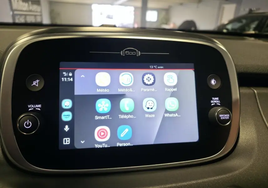 Écran tactile multimédia de la Fiat 500X 2024 affichant applications connectées, entouré de commandes volume et navigation.