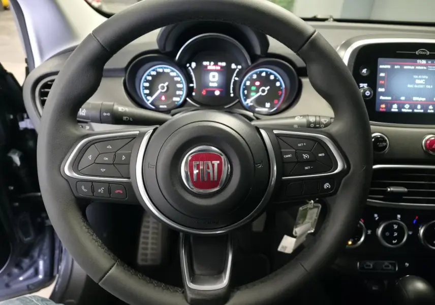 Volant en cuir noir avec logo FIAT au centre, tableau de bord digital et écran tactile du Fiat 500X gris, vue de face intérieure.