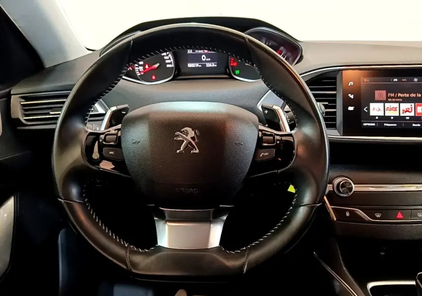 Vue intérieure centrée sur le volant cuir noir de la Peugeot 308, avec tableau de bord et écran tactile visible à droite.