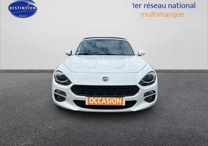 Vue de face d'une FIAT 124 Spider blanche avec phares noirs et plaque orange "OCCASION" sur un sol gris.