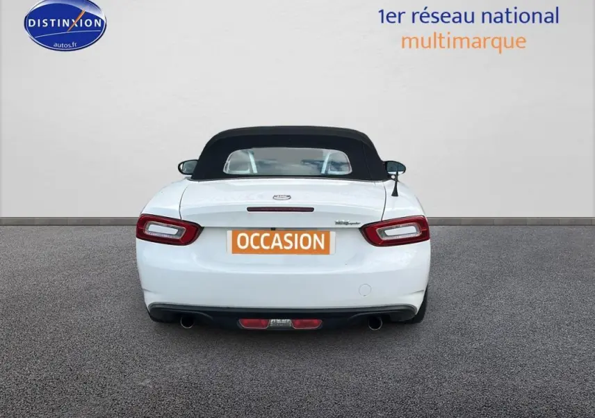 Vue arrière d'une FIAT 124 Spider blanche avec capote noire, feux arrière allongés et double sortie d'échappement.