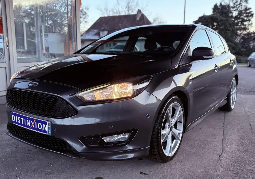 Vue 3/4 avant droite d’une Ford Focus 1.5 TDCI 120 ST Line grise avec jantes alliage 18 pouces et phares allumés.