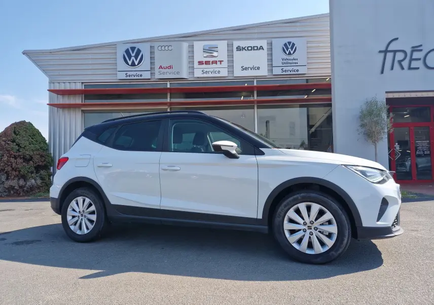 Vue de profil côté gauche d'un SEAT Arona blanc alpin avec toit noir, stationné devant un service multimarques.