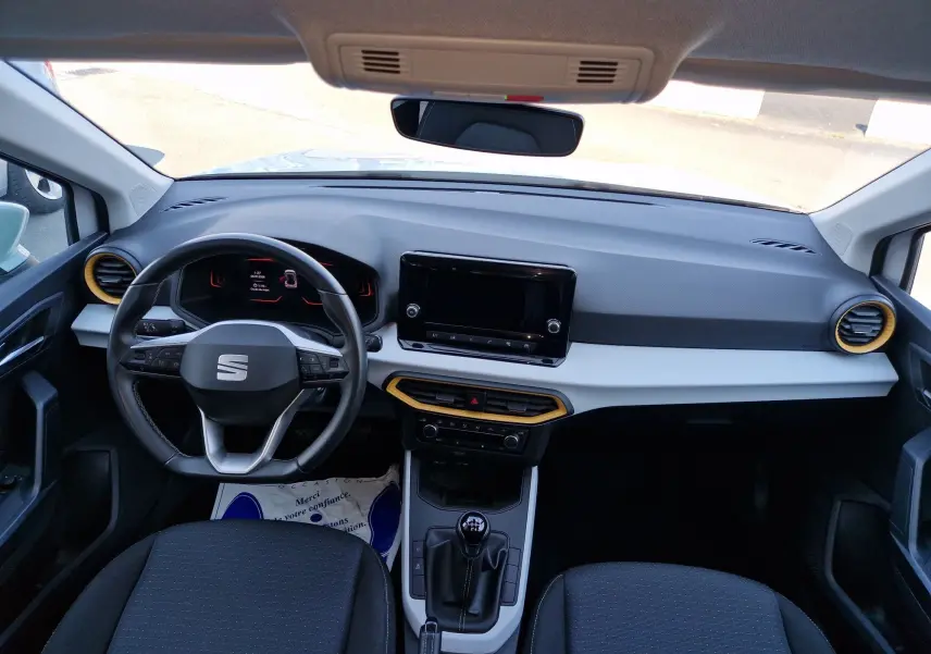 Vue intérieure avant du tableau de bord du SEAT Arona 2025, volant multifonction, console centrale avec boîte manuelle, aérateurs cerclés jaune.