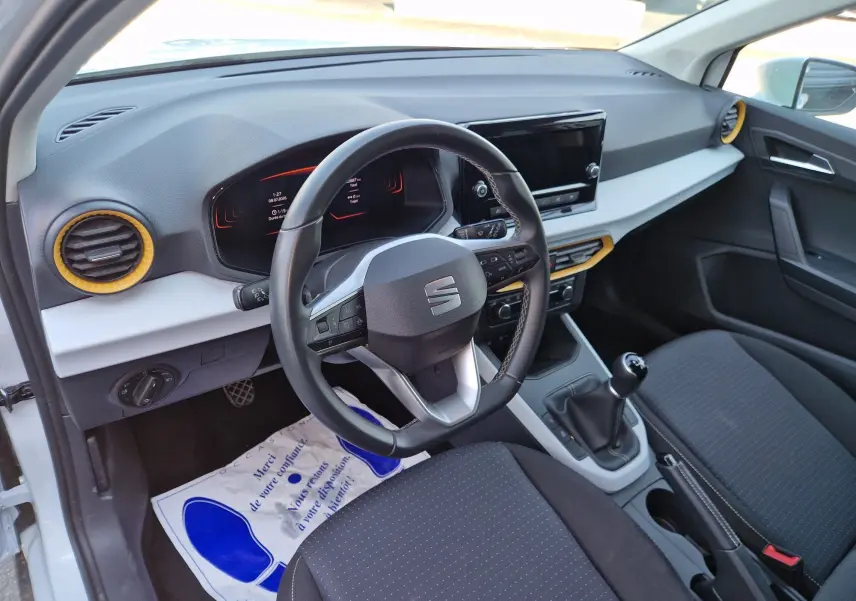 Vue intérieure côté conducteur du SEAT Arona 2025, volant cuir, tableau de bord noir avec inserts jaunes et boîte manuelle.