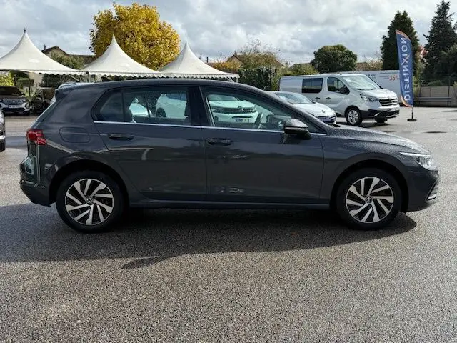 Profil côté gauche d'une Volkswagen Golf VIII 1.4 e-Hybrid gris dauphin garée en extérieur sous un ciel nuageux.