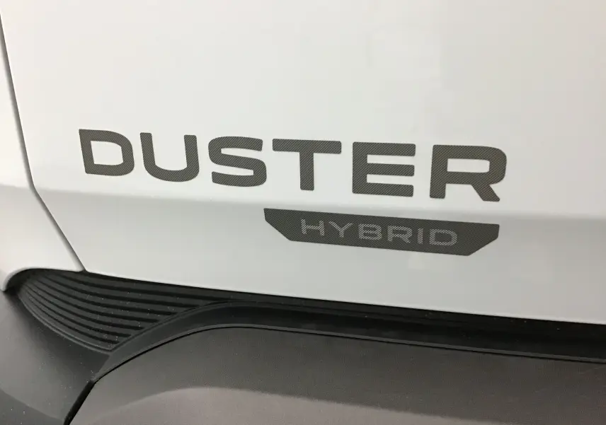 Gros plan sur le logo DUSTER Hybrid en gris sur la carrosserie blanc glacier du SUV Dacia Duster 2025.