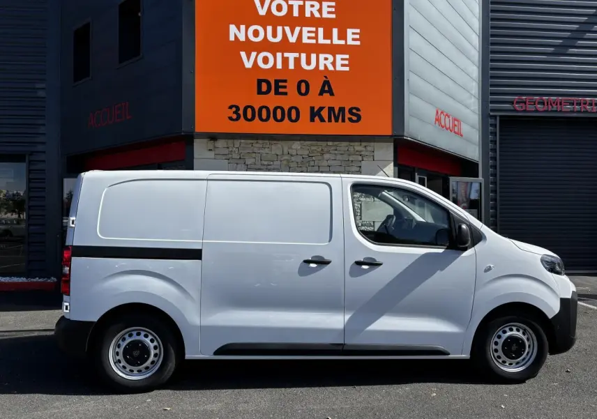 Profil droit d'un Peugeot Expert Fourgon blanc 2025 garé devant un bâtiment avec panneau orange "Votre nouvelle voiture".