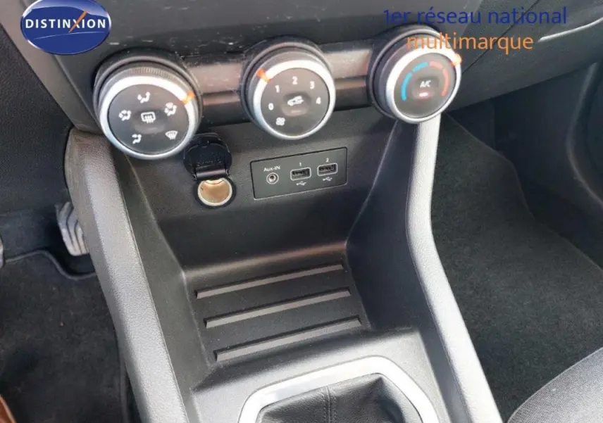 Vue intérieure centrée sur la console centrale de la Renault Clio blanche, avec commandes de climatisation et prise auxiliaire visibles.