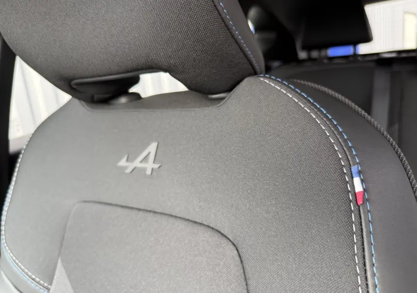 Gros plan sur l'appui-tête noir du siège avant avec logo Alpine embossé et surpiqûres bleues et drapeau tricolore.