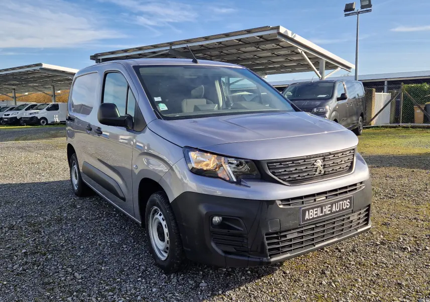 Peugeot Partner gris Artense en 3/4 avant droit, utilitaire compact avec calandre noire et phares allumés sur parking extérieur.