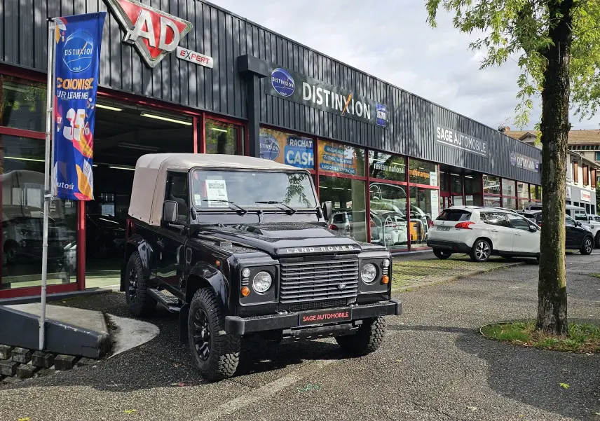 Land Rover 90 Defender noir 3/4 avant avec toit souple beige, stationné devant un garage automobile.