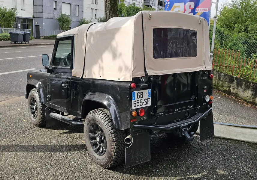 Land Rover 90 Defender noir vu en 3/4 arrière droit avec capote souple beige et roues tout-terrain.