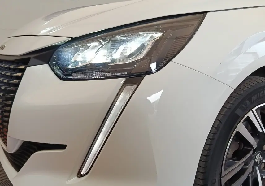 Gros plan sur le phare avant droit allumé d'une Peugeot 208 blanche, avec partie de la calandre et roue visible.