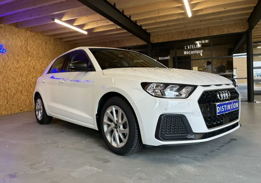 Audi A1 blanc vue 3/4 avant droit, avec jantes alu 17 pouces et calandre noire en intérieur d'atelier.