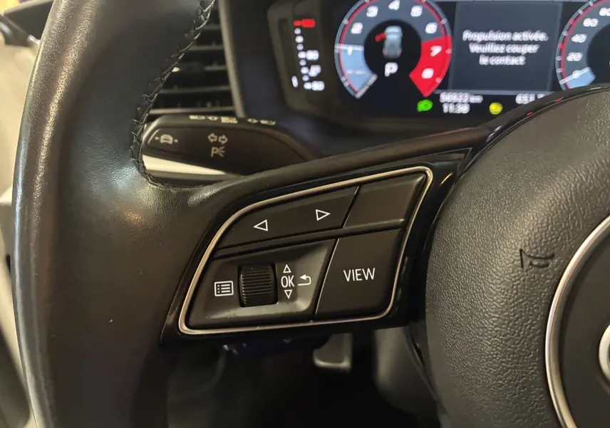 Gros plan sur les commandes gauche du volant cuir noir d’une Audi A1 blanche, avec affichage numérique en arrière-plan.