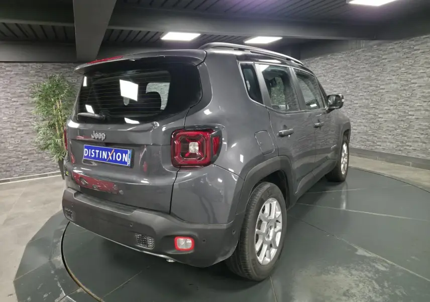 Jeep Renegade gris métallisé vu en 3/4 arrière droit dans un showroom avec toit noir et barres de toit.