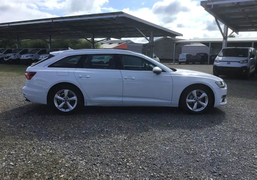 Audi A6 Avant blanc glacier vue de profil côté gauche sur un sol gravillonné avec abris de parking en arrière-plan.