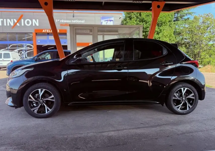 Profil côté gauche d'une Toyota Yaris Hybrid noire 2025, soulignant ses jantes alliage et lignes compactes sous un abri.