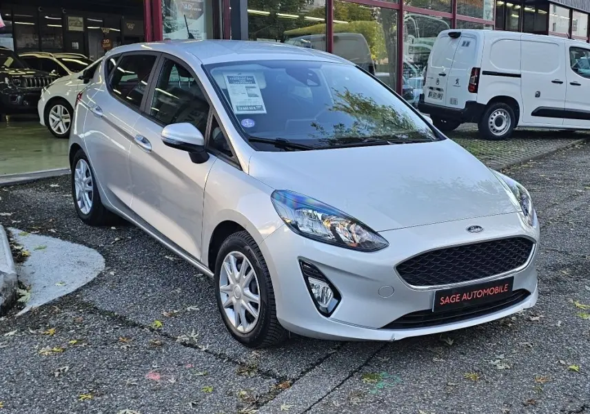 Ford Fiesta 2021 gris lunaire métal vue 3/4 avant droit, avec calandre noire et phares allumés devant un garage Sage Automobile.