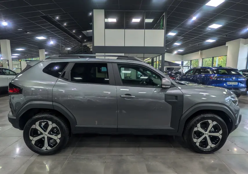 Profil côté droit du Dacia Duster gris schiste 2025 en version Mild Hybrid 130 CH 4x4, avec jantes bi-ton et barres de toit.