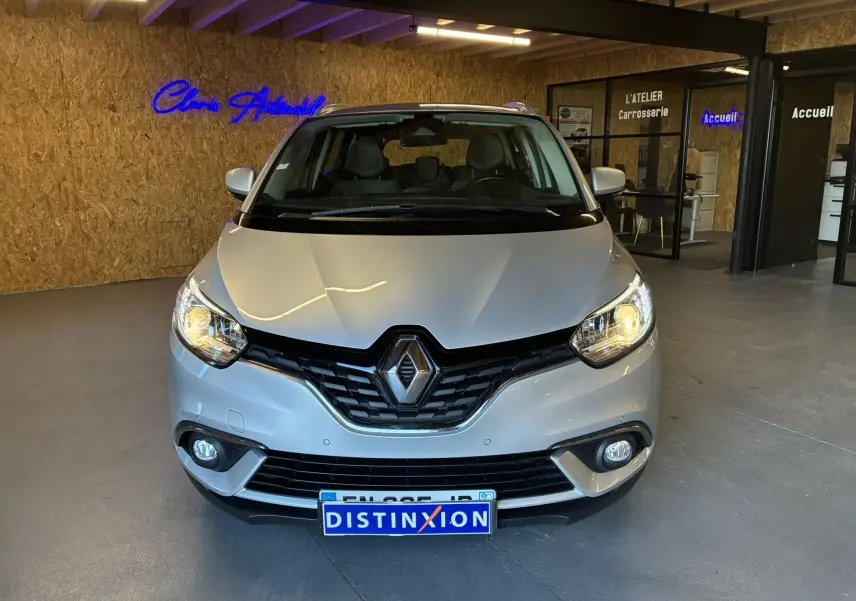 Vue de face d'un Renault Grand Scenic gris 2017 avec phares allumés dans un showroom moderne.