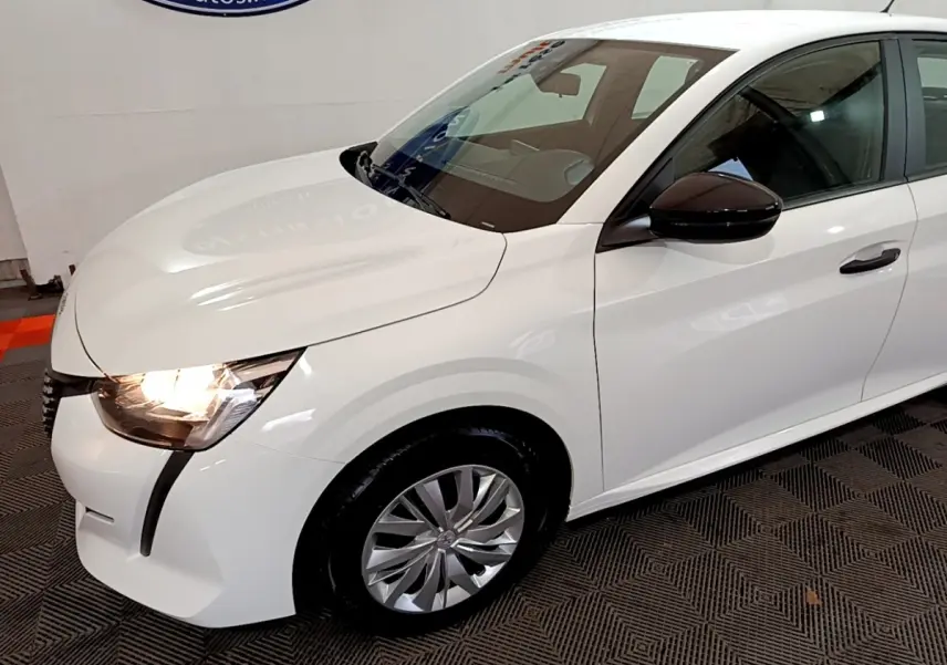 Vue 3/4 avant droit d'une Peugeot 208 blanche avec phares allumés et rétroviseur noir brillant.