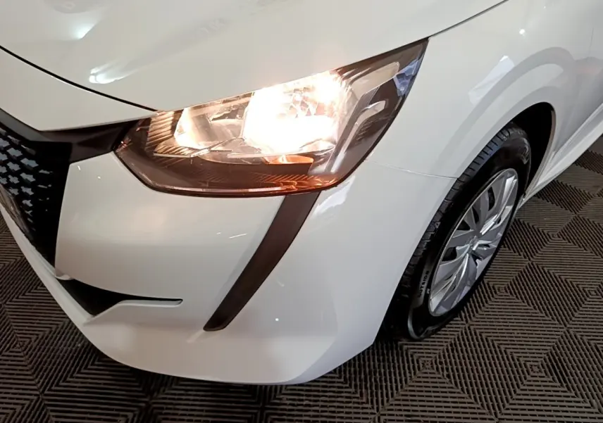 Gros plan sur l'avant droit blanc d'une Peugeot 208 2022 avec phare allumé et jante visible sur sol texturé.