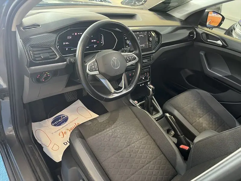 Vue intérieure côté conducteur du Volkswagen T-Cross gris, mettant en valeur le tableau de bord digital et les sièges en tissu noir à motifs.