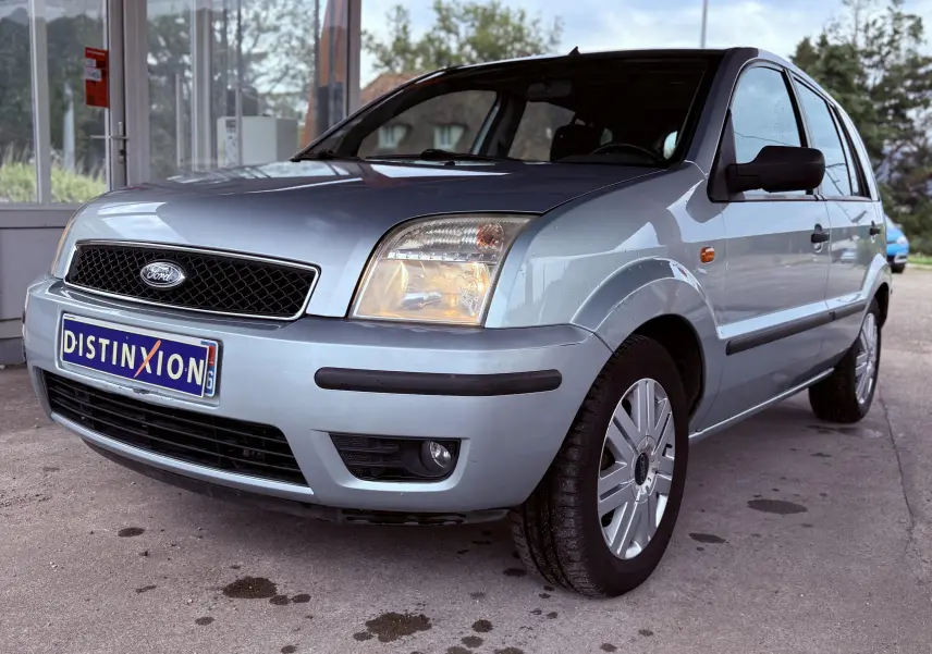 Ford Fusion bleu clair vue 3/4 avant droit avec phares allumés et plaque d'immatriculation Distinxion visible.