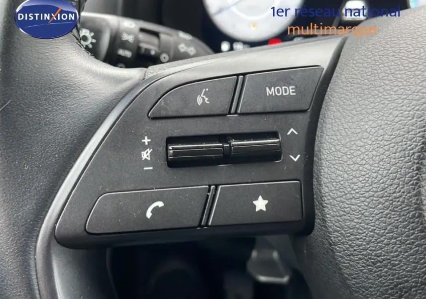 Gros plan sur les commandes gauche du volant noir de la Hyundai i20 2025, boutons mode, volume et téléphone visibles.