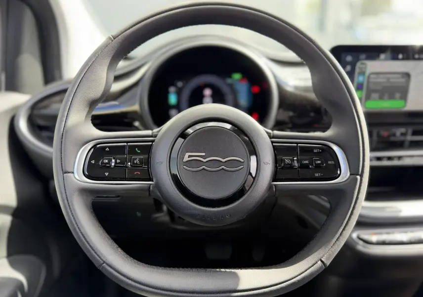 Vue rapprochée du volant noir Soft-Touch du Fiat 500 électrique 2024 avec commandes intégrées et tableau de bord numérique flou.
