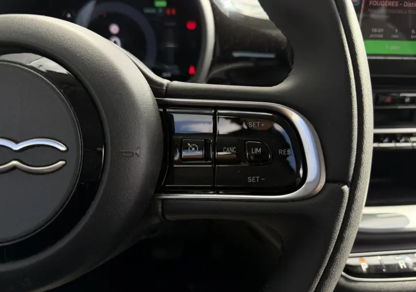 Gros plan sur le volant noir Soft-Touch de la Fiat 500 électrique 2024, avec commandes de régulateur de vitesse visibles.