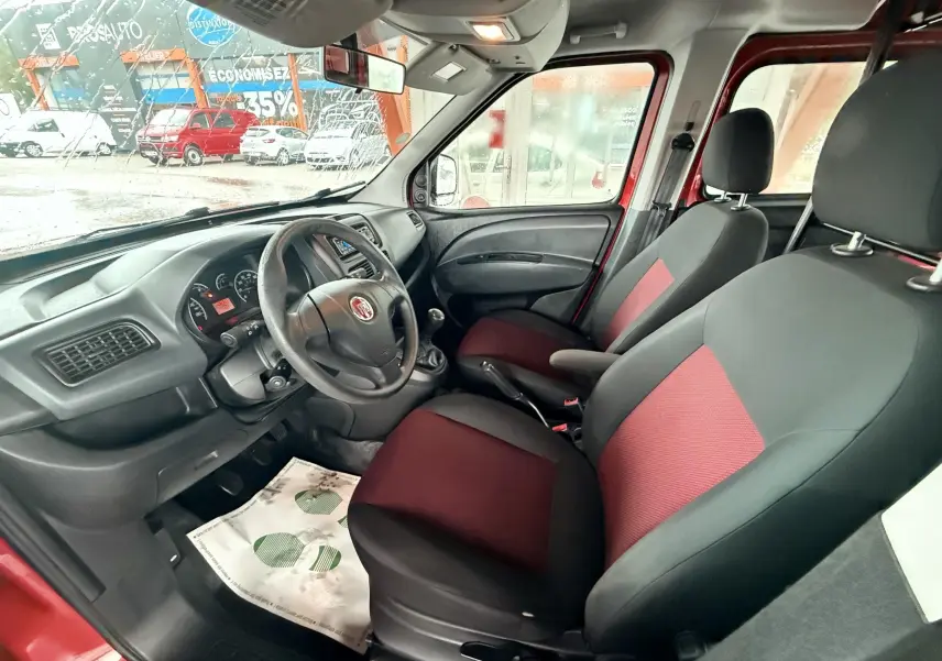 Vue intérieure côté conducteur du Fiat Doblo Cargo 2011, sièges tissu rouge et noir, volant cuir et tableau de bord noir.
