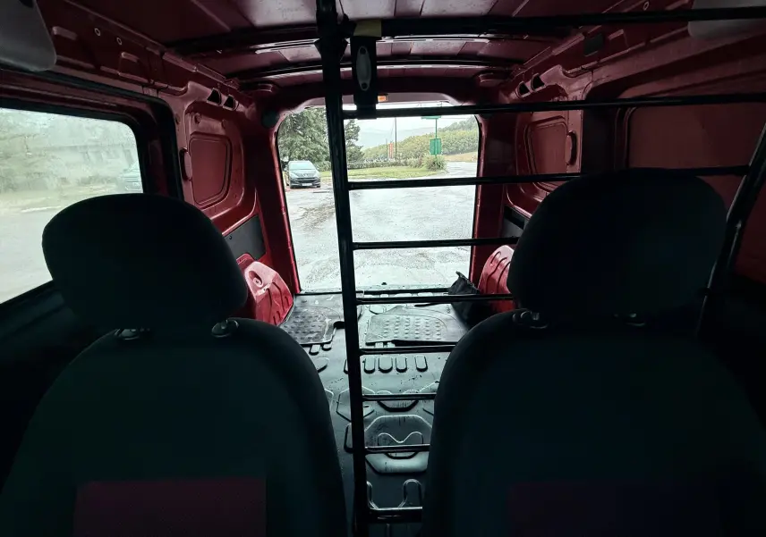 Vue intérieure arrière du Fiat Doblo Cargo rouge, montrant les sièges avant et la cloison métallique séparant l'espace de chargement.