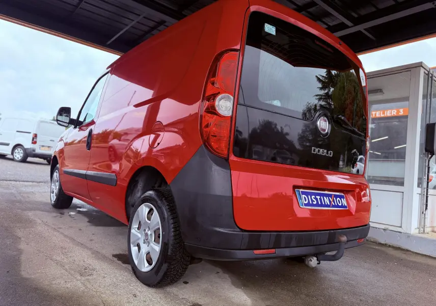 Vue 3/4 arrière droite d’un Fiat Doblo Cargo rouge avec hayon noir et attelage, stationné sous un abri.