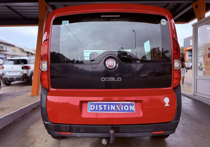 Vue arrière d’un FIAT Doblo Cargo rouge avec hayon noir et attelage, stationné sous un abri.