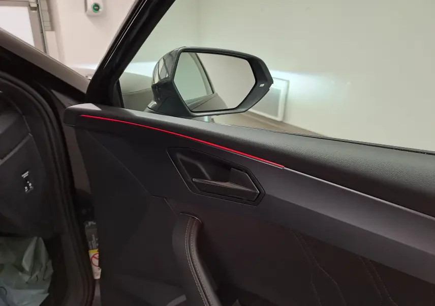 Intérieur côté conducteur de la CUPRA Leon 2025 noir minuit, avec éclairage d'ambiance rouge sur la porte et miroir latéral visible.