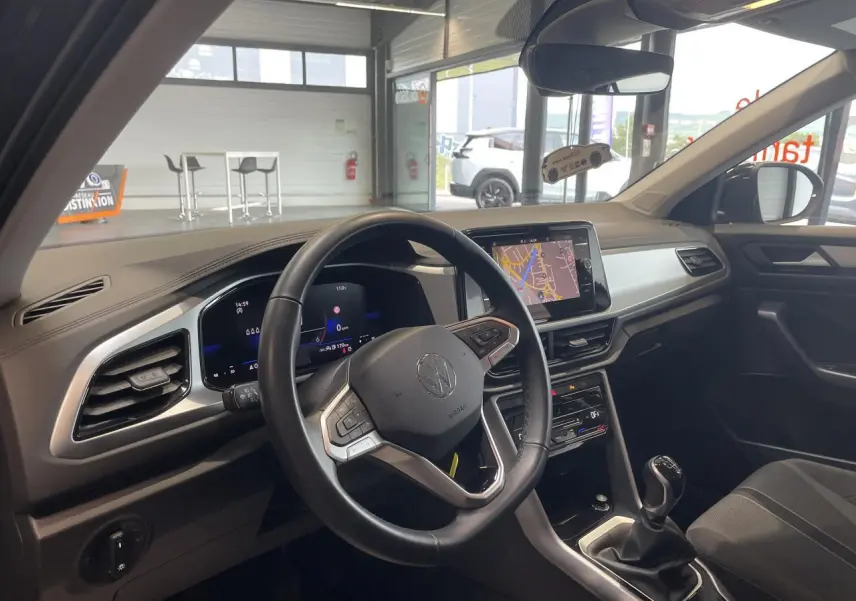 Vue intérieure côté conducteur du Volkswagen T-Roc 2025 gris Indium, avec volant cuir multifonction et écran tactile central.