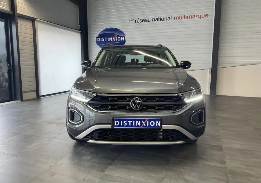 Vue frontale d'un Volkswagen T-Roc 2025 gris Indium avec calandre noire et éclairage LED allumé en intérieur showroom.