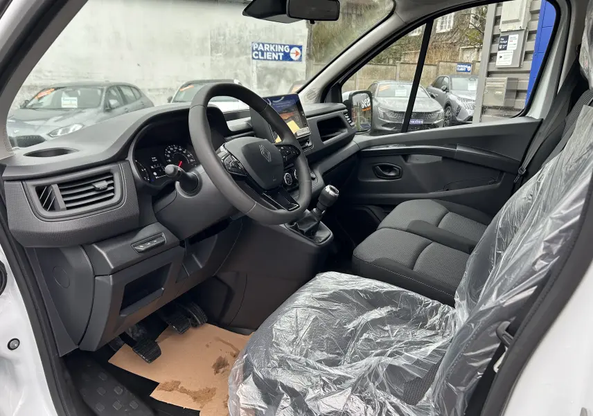 Vue intérieure côté conducteur du Renault Trafic Combi blanc Glacier 2025, montrant le volant, la console et les sièges en tissu protégés.