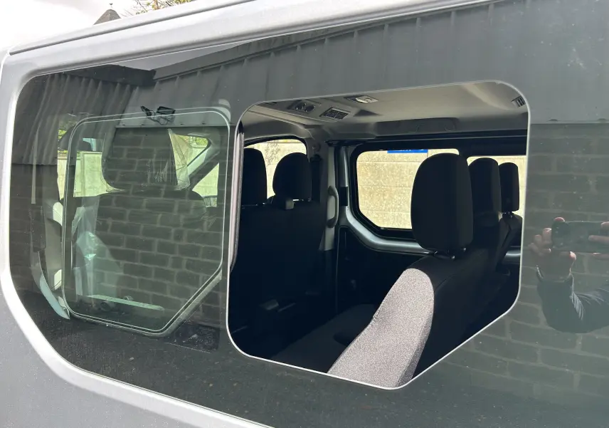 Vue intérieure par la porte latérale gauche ouverte du Renault Trafic Combi blanc glacier, montrant les sièges en tissu noir.