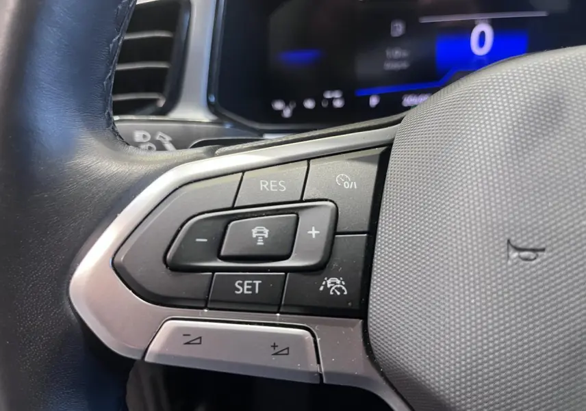 Gros plan sur les commandes au volant à gauche du Volkswagen T-Roc 2025, avec affichage digital bleu en arrière-plan.