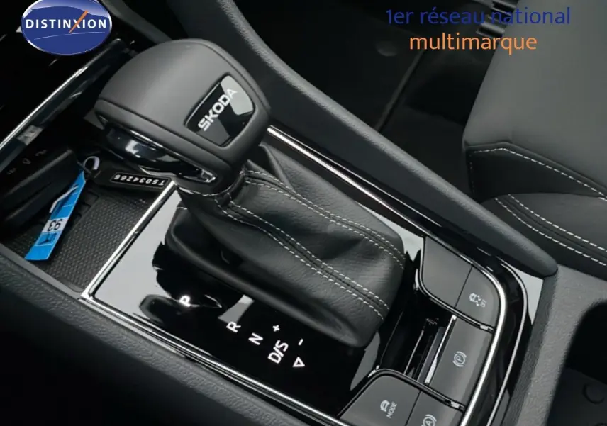 Gros plan sur le levier de vitesse automatique noir avec logo SKODA, entouré de commandes et coutures blanches visibles.